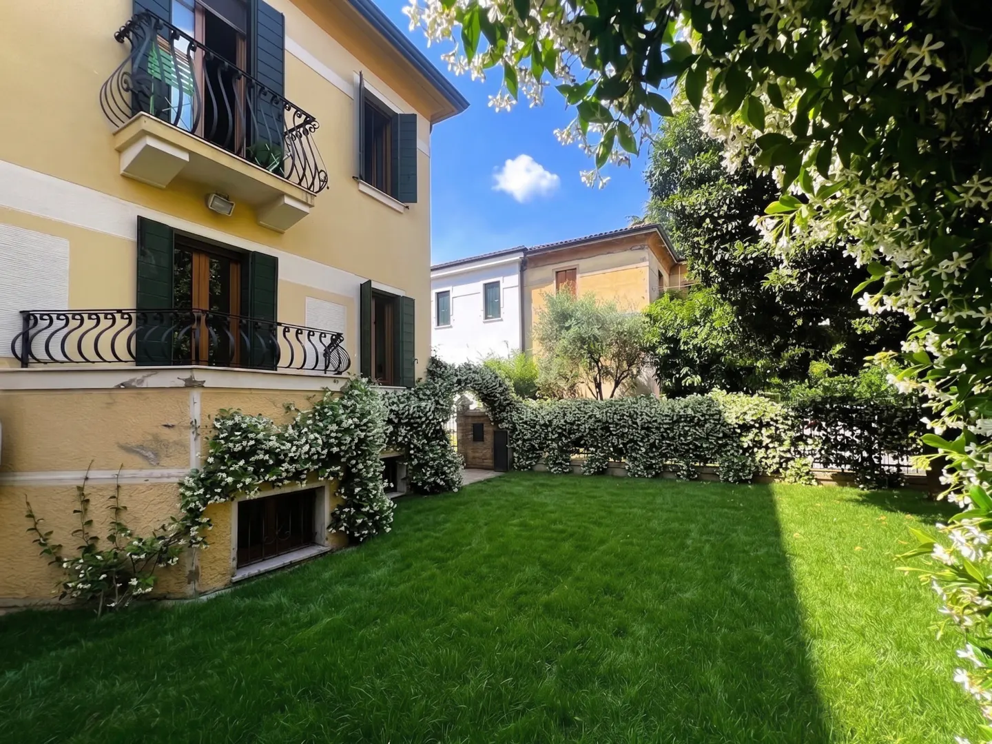 A yellow house with green shutters and black wrought iron balconies overlooks a green lawn and a hedge archway.