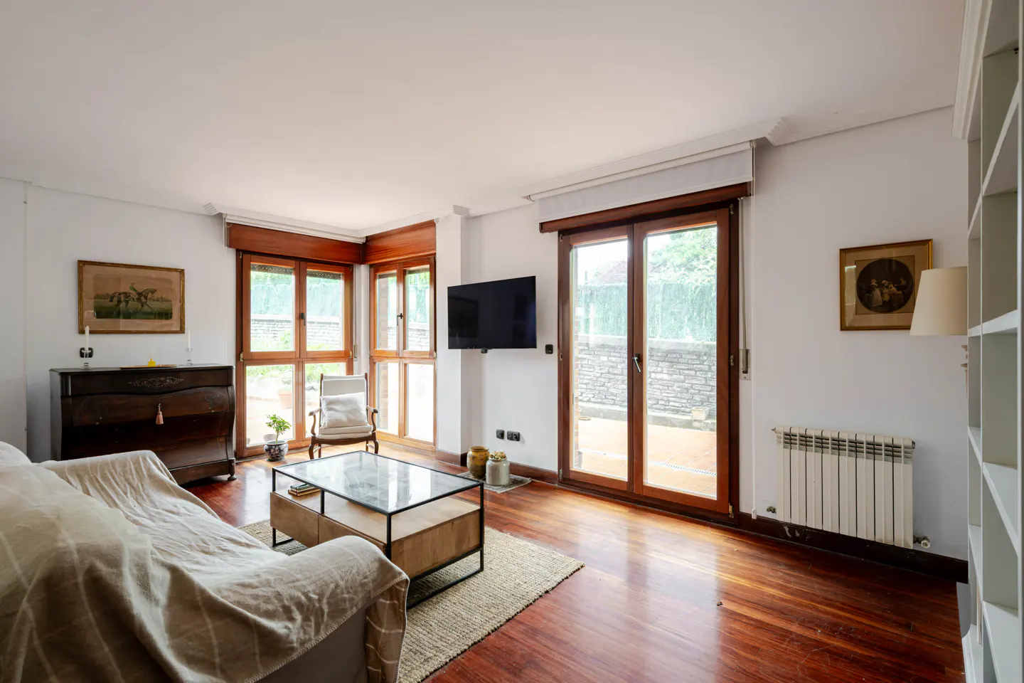 Bright living room with hardwood floors, a beige sofa, glass coffee table, wall-mounted TV, and glass doors to a patio.