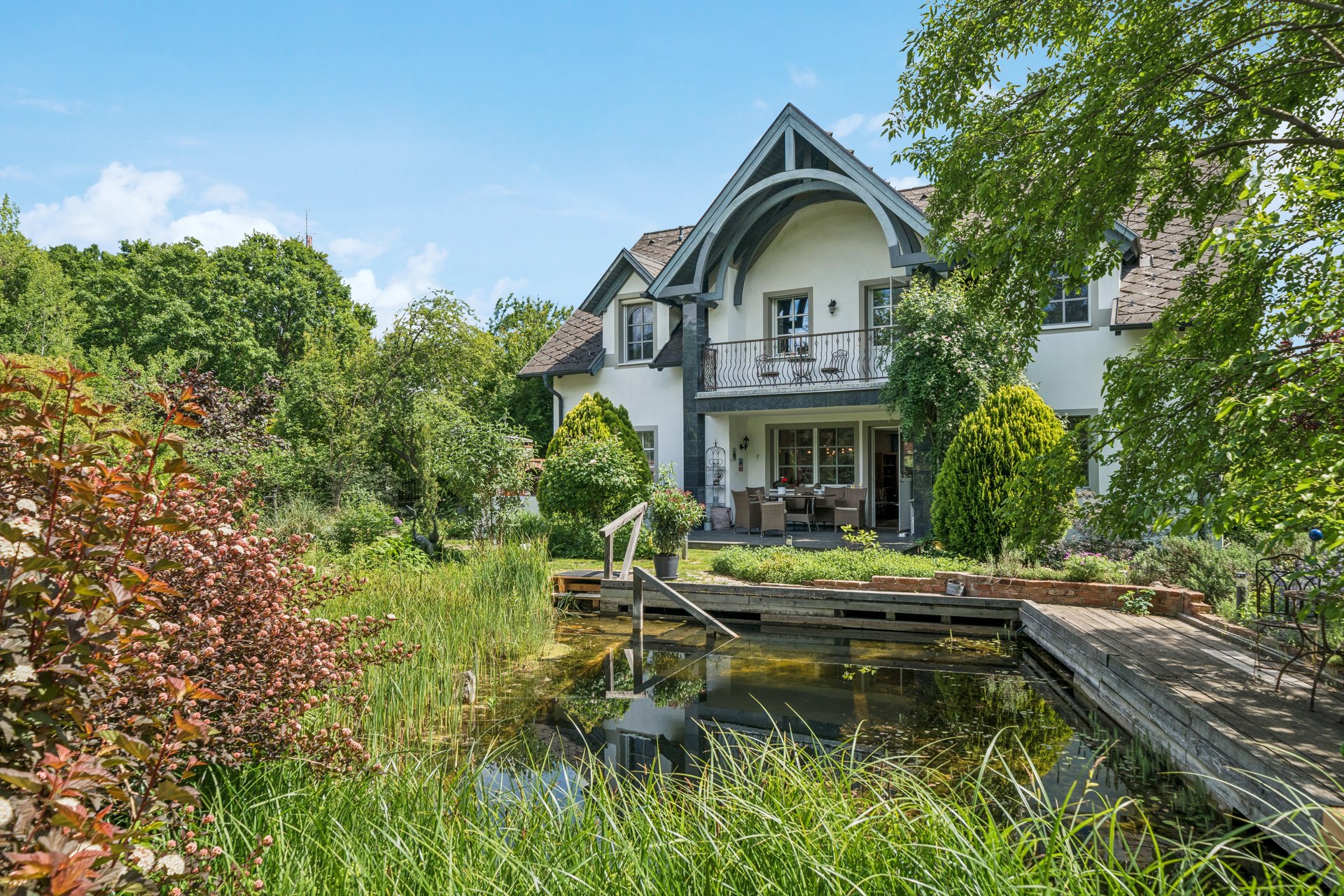 A white two-story house with a balcony and patio sits behind a lush garden pond with a wooden deck and tall grasses.