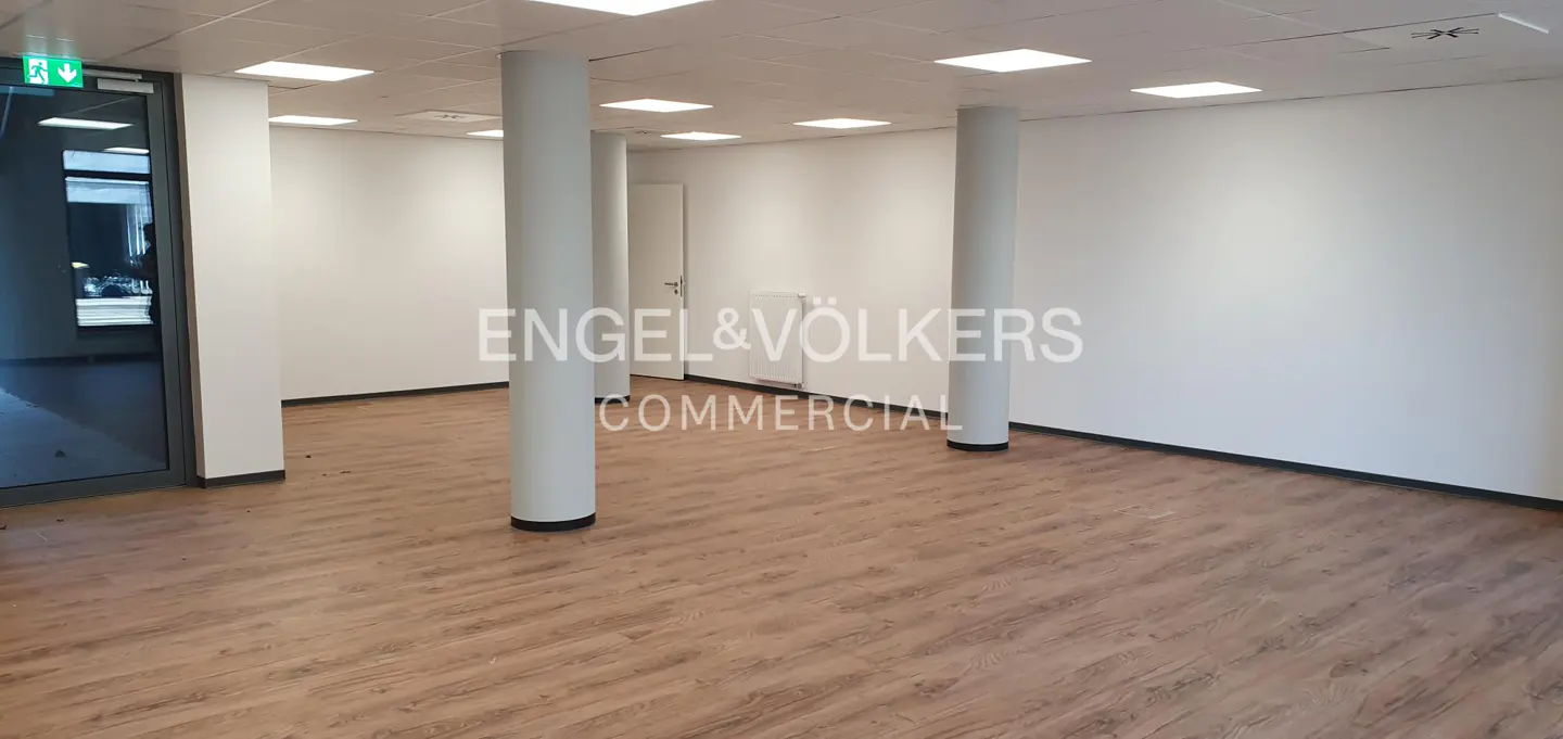 Empty commercial space with wood floors, white walls, and support columns. Engel & Völkers logo in the center.