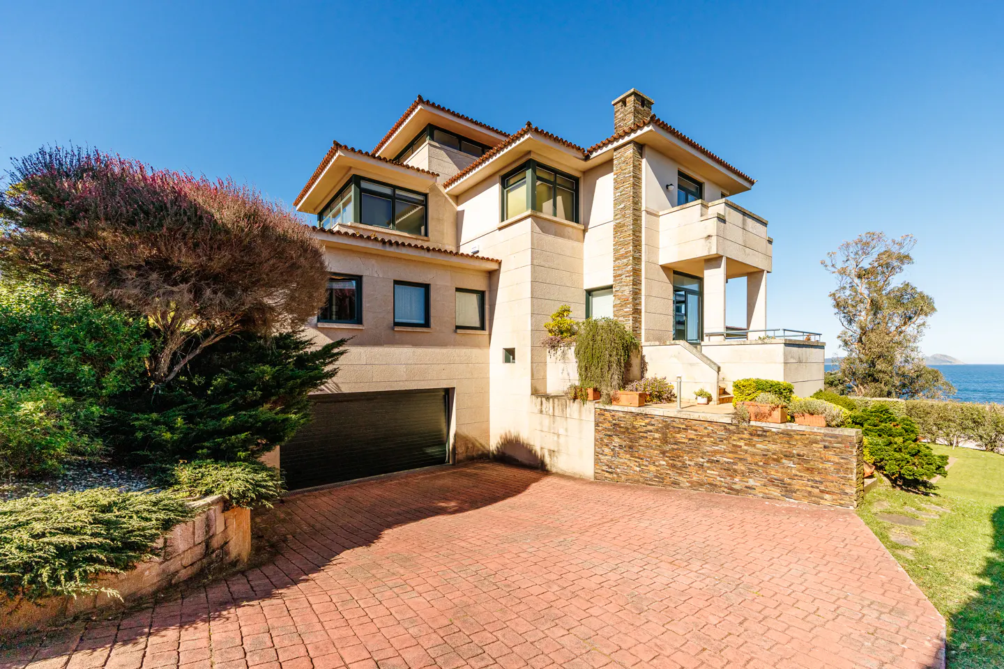 A modern, multi-story beige house with a red brick driveway and a blue sky background.