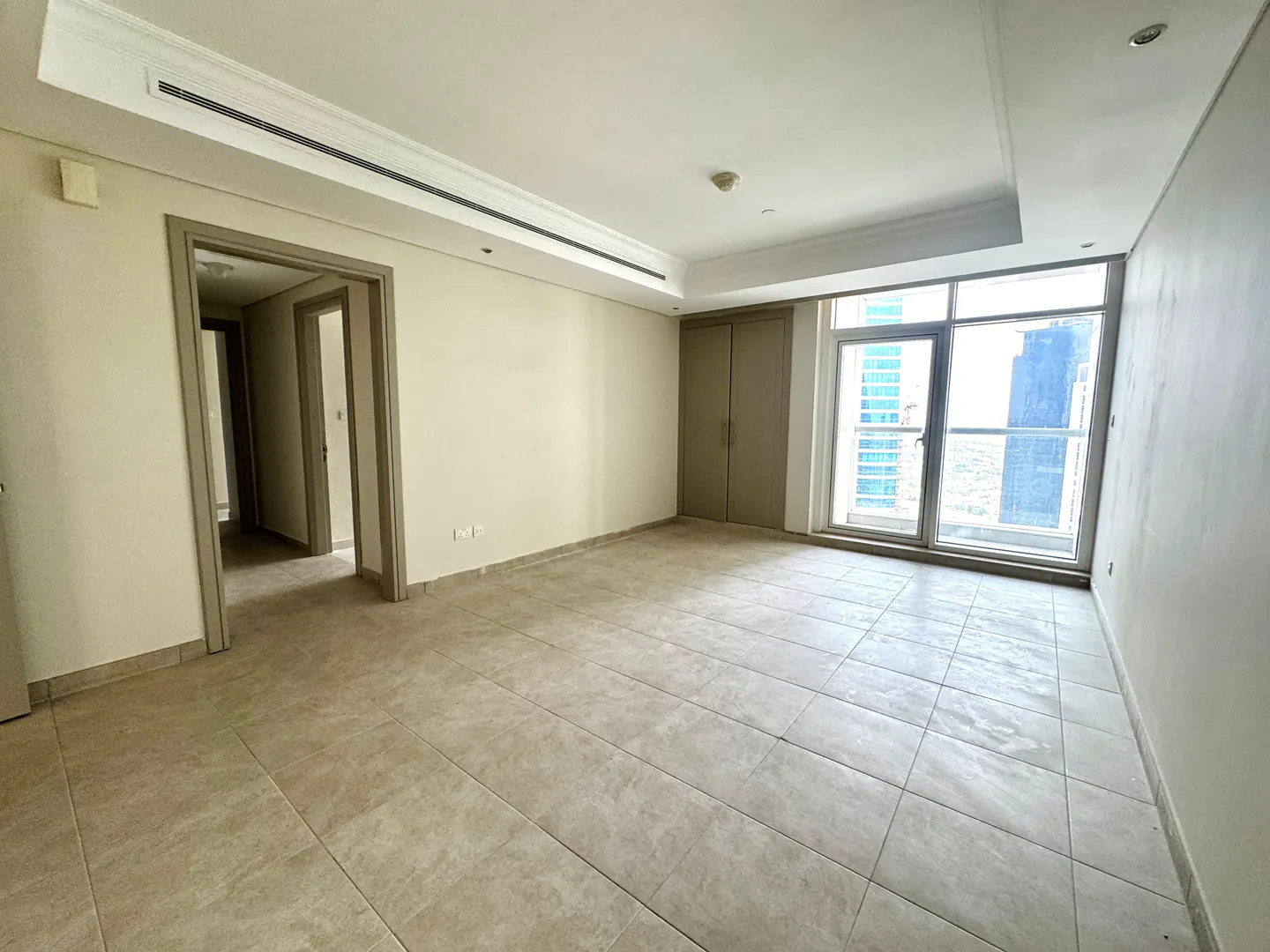 An empty room with beige tile flooring, white walls, and a large window with a city view. Two doorways are visible.