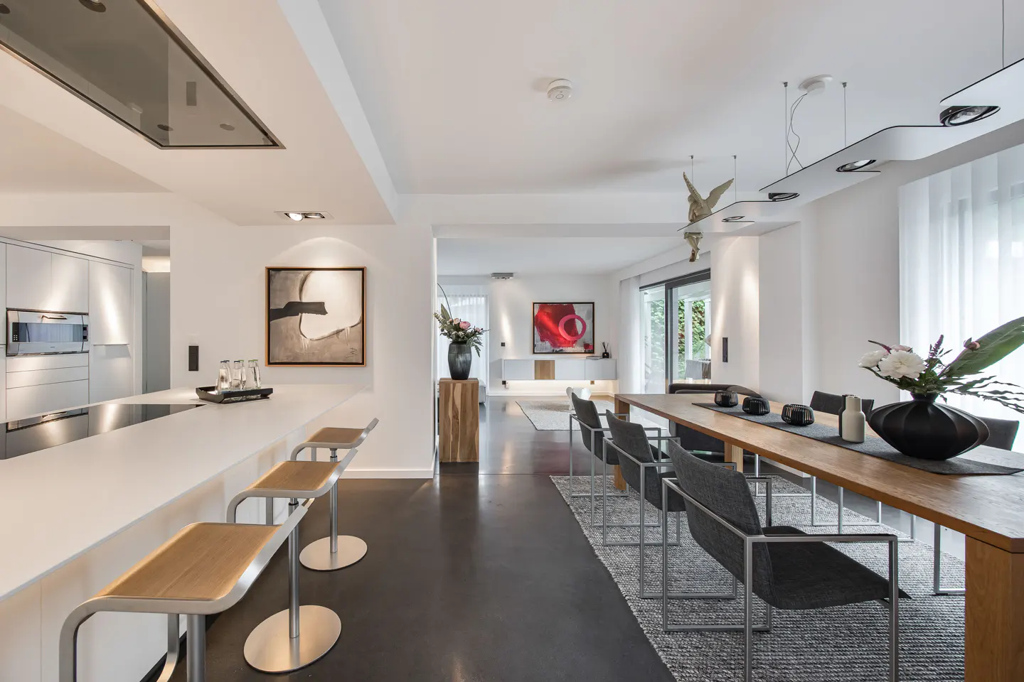Modern open-plan kitchen and dining area with white walls, dark floors, a wooden dining table, and contemporary bar stools.