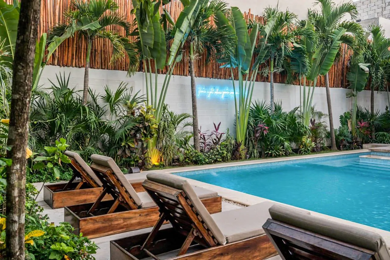 A turquoise pool is surrounded by lounge chairs and lush tropical plants. A white wall and bamboo fence are in the background.