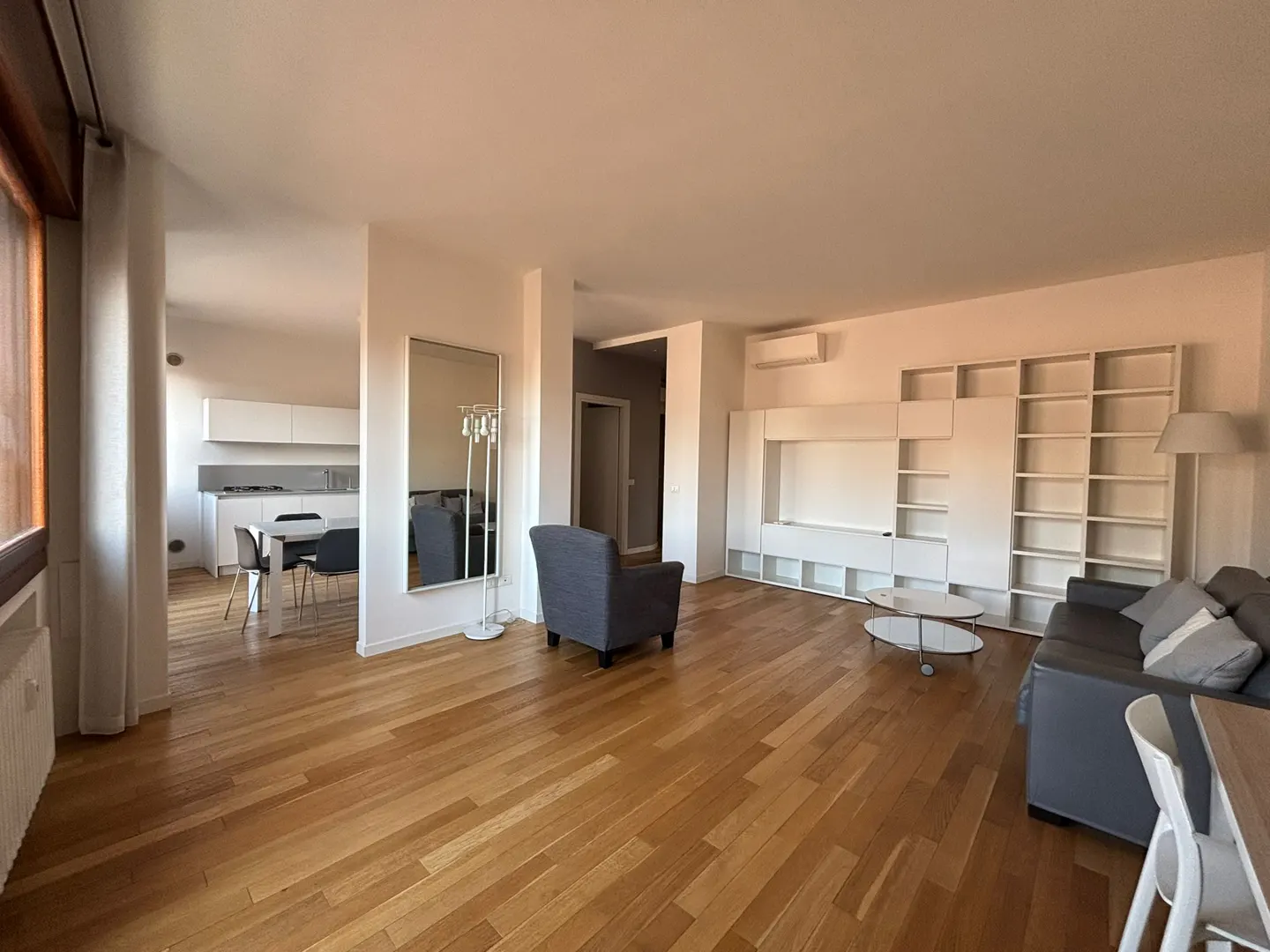 Bright, open-concept living space with hardwood floors, a gray sofa, armchair, and a white built-in shelving unit. A kitchen is visible in the background.