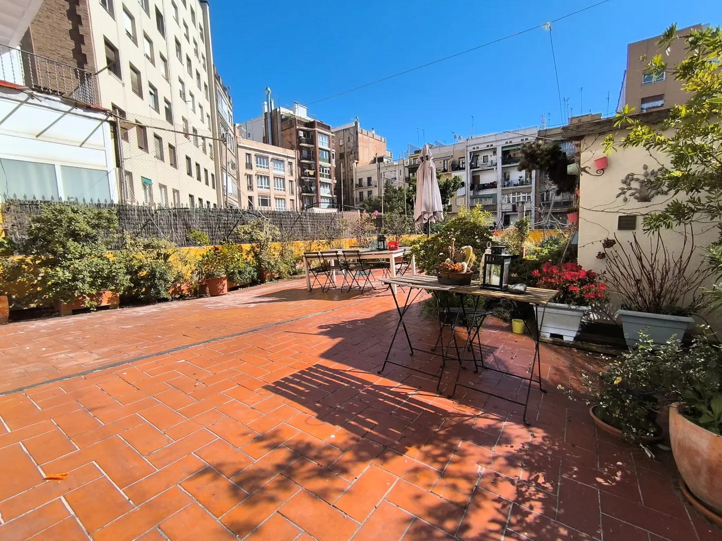 Outdoor patio with red brick flooring, tables, chairs, and potted plants. Buildings are visible in the background under a clear blue sky.