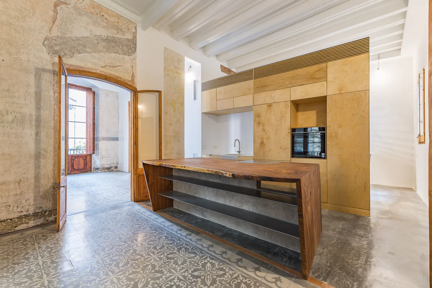 Interior view of a kitchen with a wooden island, cabinets, and patterned tile flooring. An open doorway leads to another room with a window.