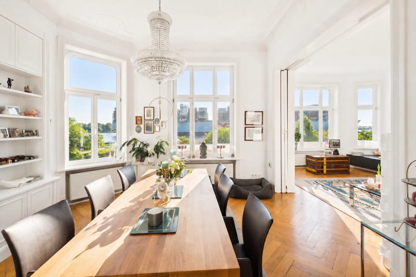 Bright dining room with a long wooden table, black chairs, and a crystal chandelier. Large windows offer a view of trees and water.