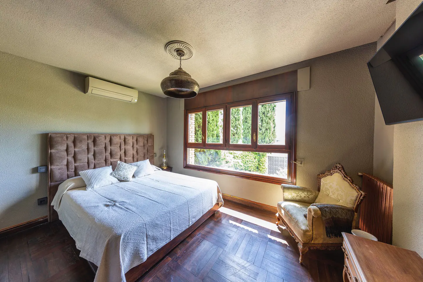 Bedroom with a brown headboard bed, white linens, and a vintage chair by a window with green trees outside.