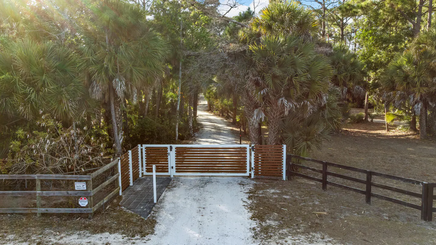 Gated driveway entrance to a property. White and brown horizontal slat gate with a sandy drive lined with palm trees.