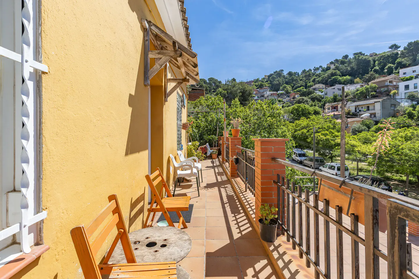 Balcony view with orange chairs, a wooden table, and a yellow wall. A metal railing overlooks a green hillside with houses.