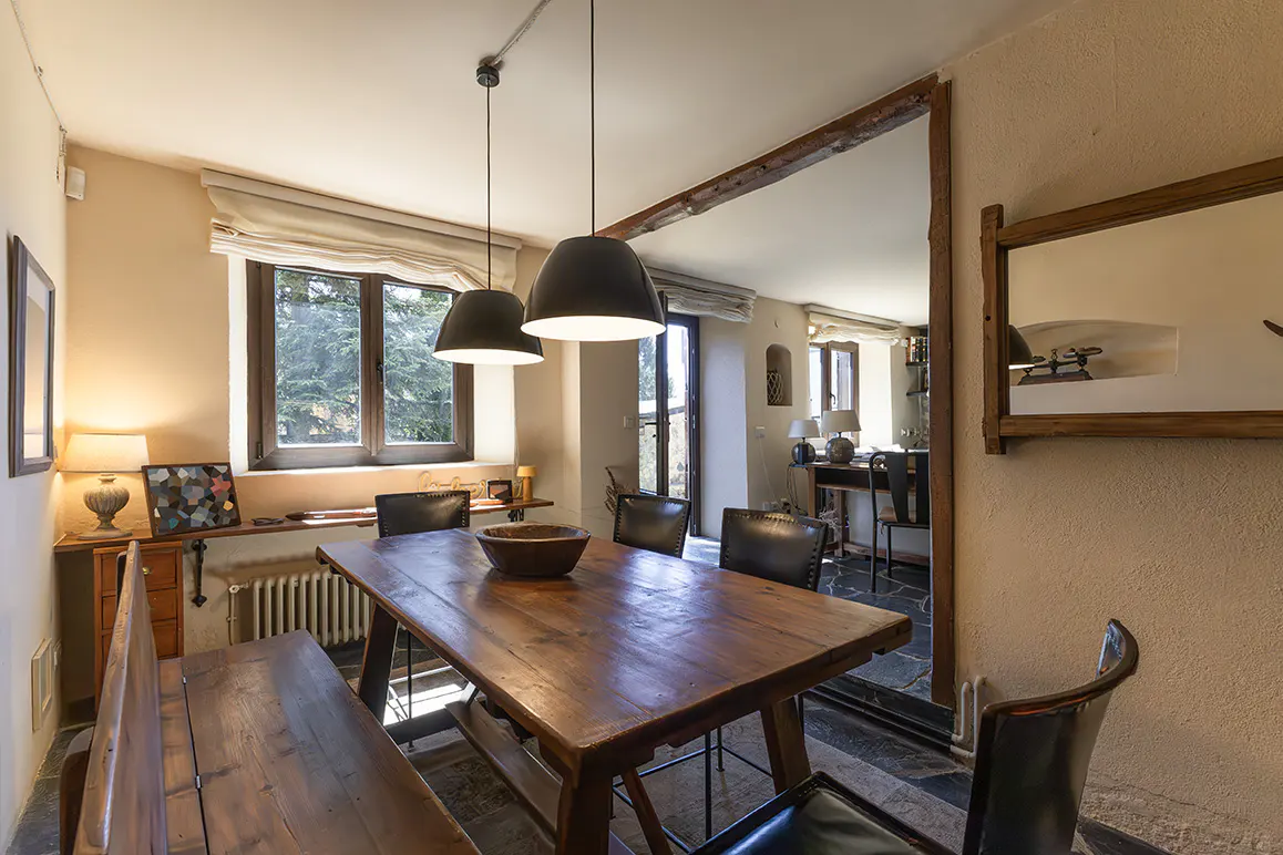 Rustic dining room with a wooden table, bench, and black leather chairs under two black pendant lights. A window and mirror add light.