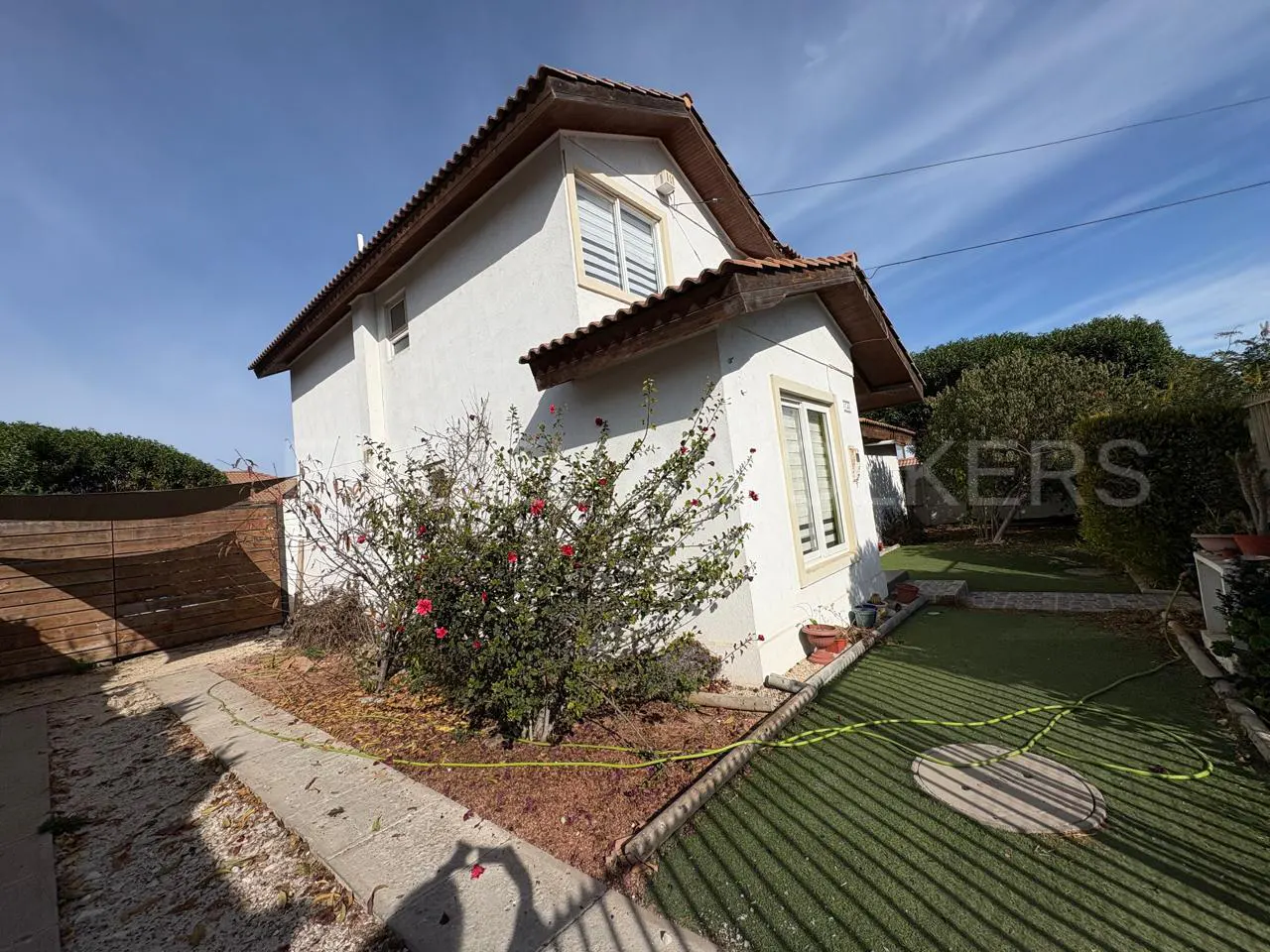 White two-story house with a brown roof, a flowering hibiscus bush, and a small green lawn under a bright blue sky.