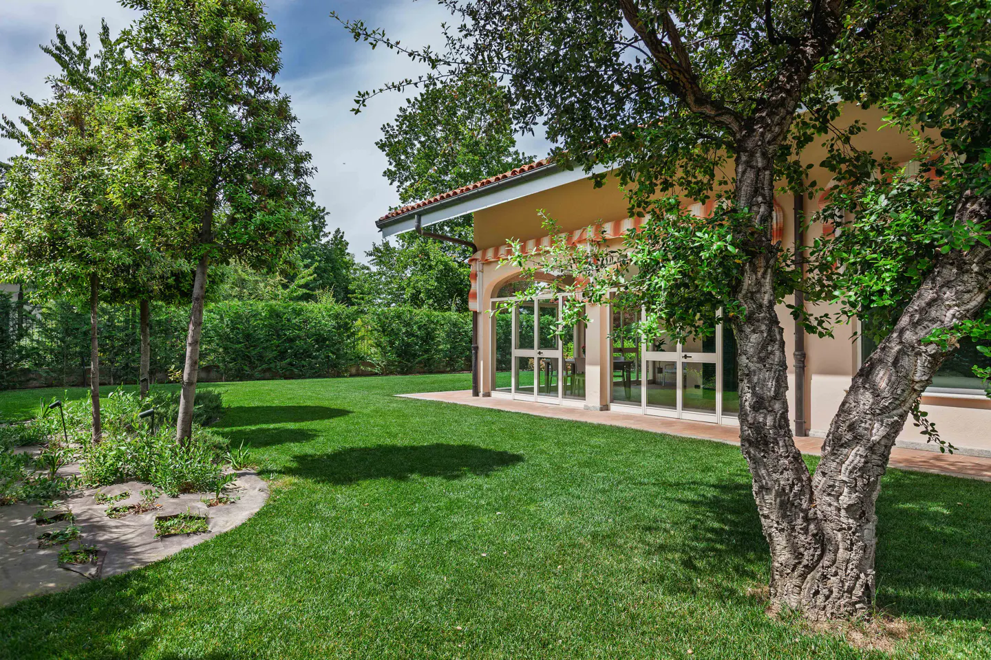 A lush green lawn with trees and a beige house featuring a glass-enclosed patio under a striped awning.