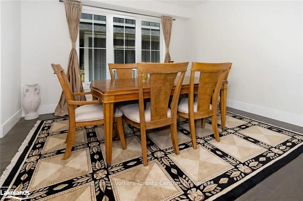 Dining room with a wooden table and chairs on a patterned rug. A white vase sits near a window with beige curtains.