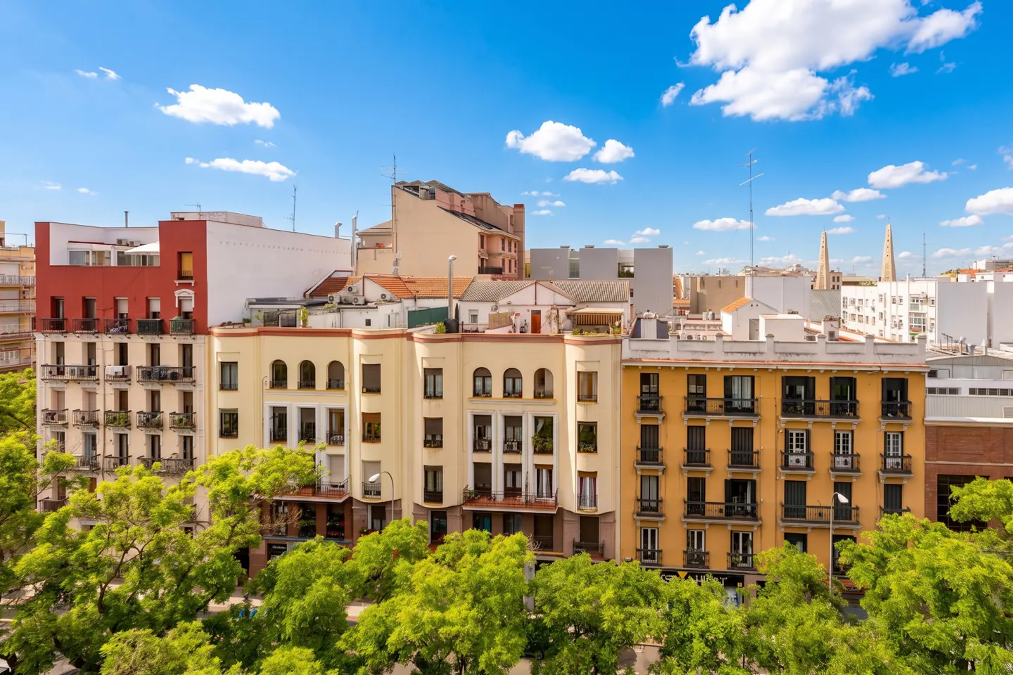 Cityscape view of colorful buildings with green trees in the foreground under a blue sky with white clouds.