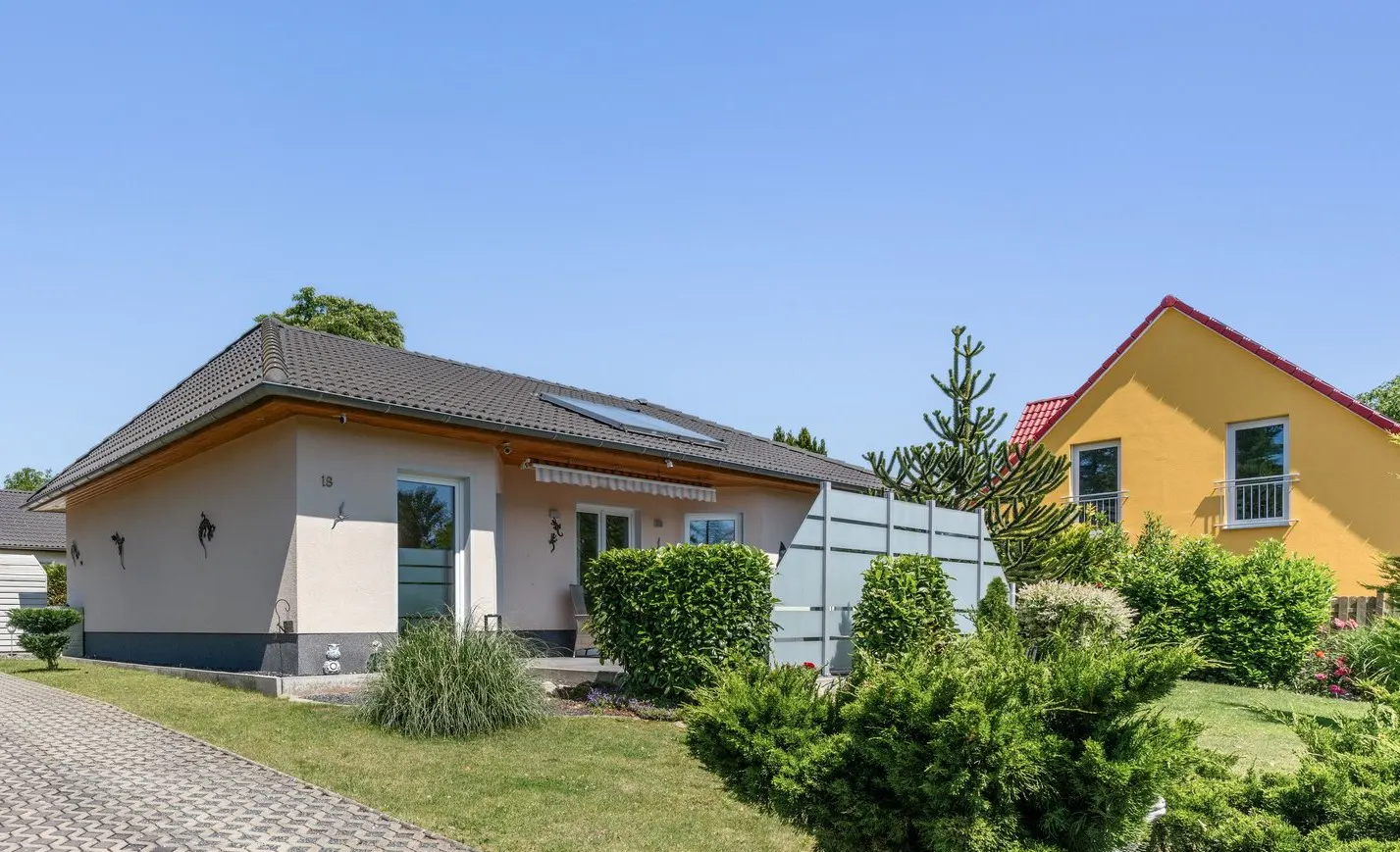 A single-story beige house with a dark roof, green lawn, and a yellow house in the background.