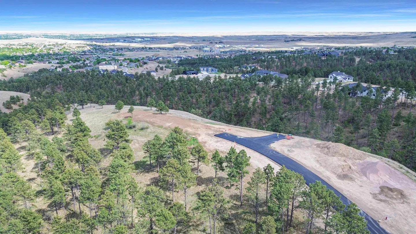 Aerial view of a newly paved T-intersection road winding through a pine forest with distant suburban homes and hills.