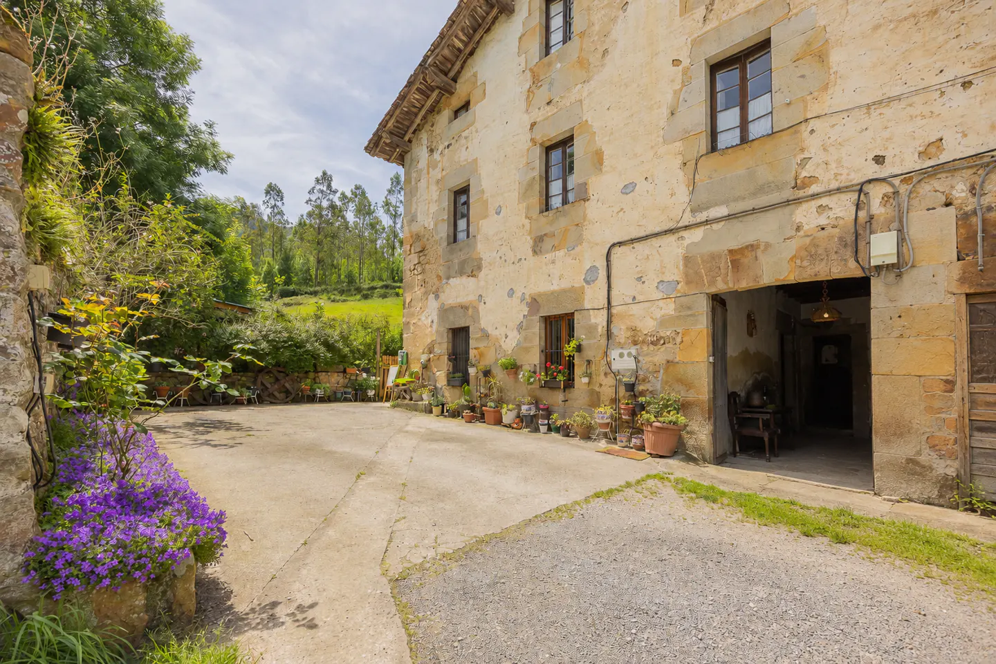 Exterior view of a stone farmhouse with flower boxes, potted plants, and a large open garage door. Purple flowers line the driveway.