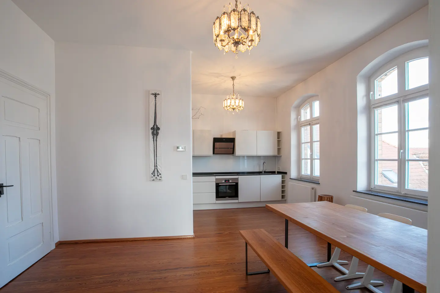 Bright, open-concept apartment with hardwood floors, white walls, and a modern kitchen. A long wooden table sits under an ornate chandelier.