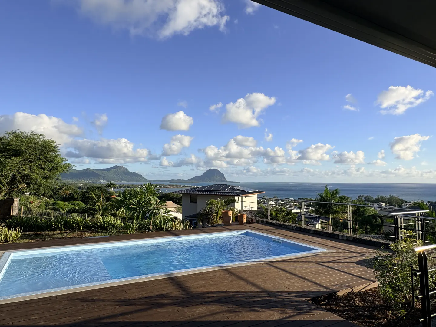 A blue swimming pool on a wooden deck overlooks a tropical landscape, ocean, and mountains under a blue, cloudy sky.