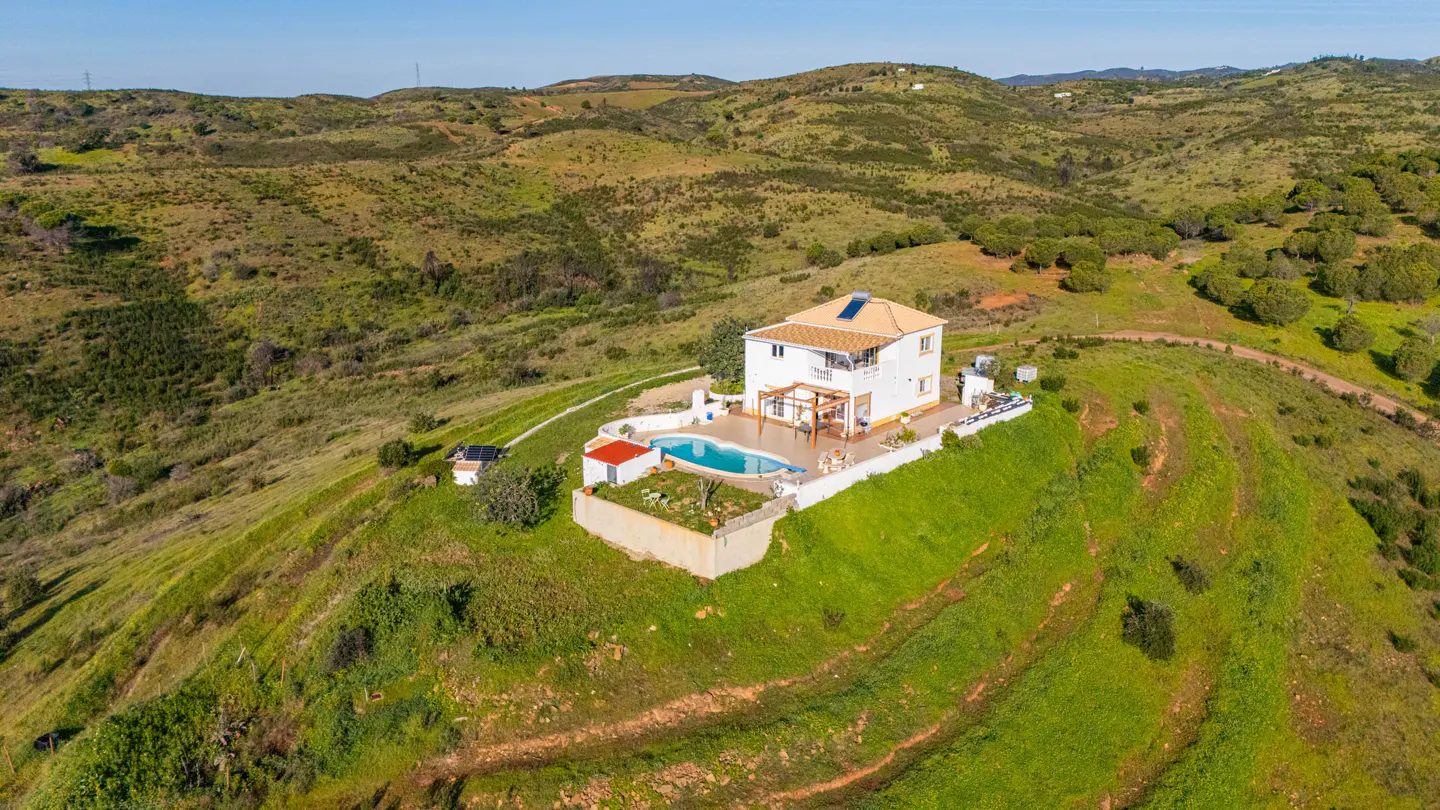 Aerial view of a white, two-story house with a pool on a green hilltop, surrounded by rolling hills under a clear blue sky.