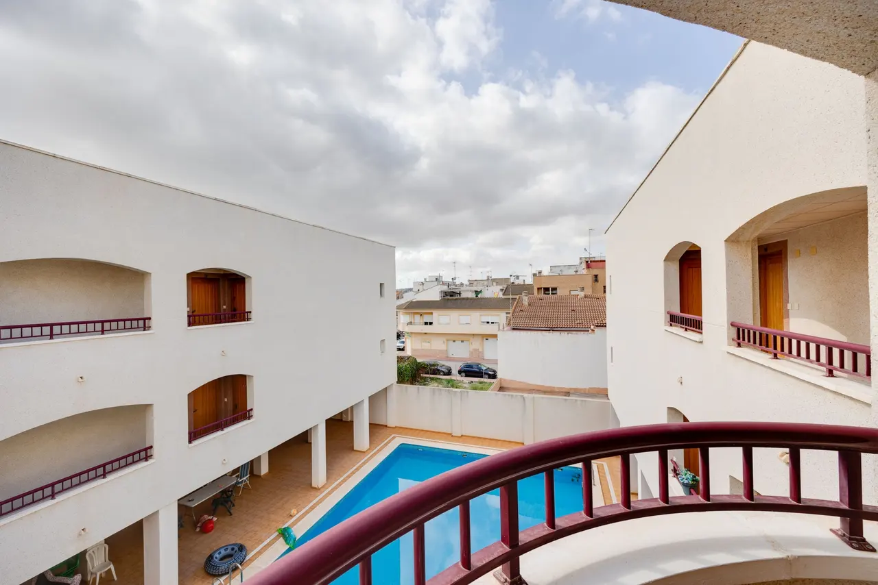 View from a balcony overlooking a blue swimming pool surrounded by white buildings under a cloudy sky.