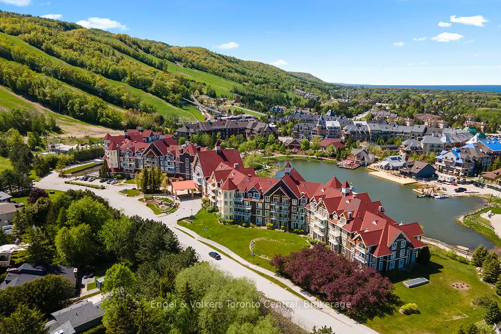 Aerial view of a resort with red-roofed buildings, a lake, and a green, forested hillside under a blue sky.