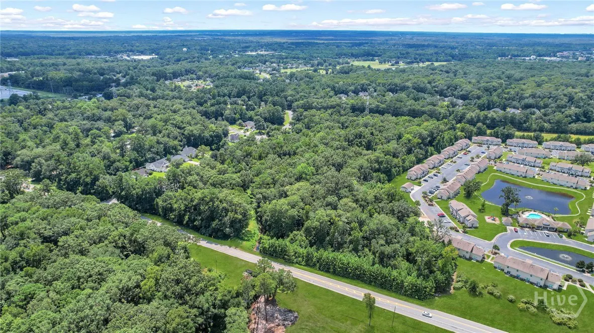 Aerial view of a wooded lot next to a road and a residential area with a pond on a sunny day.