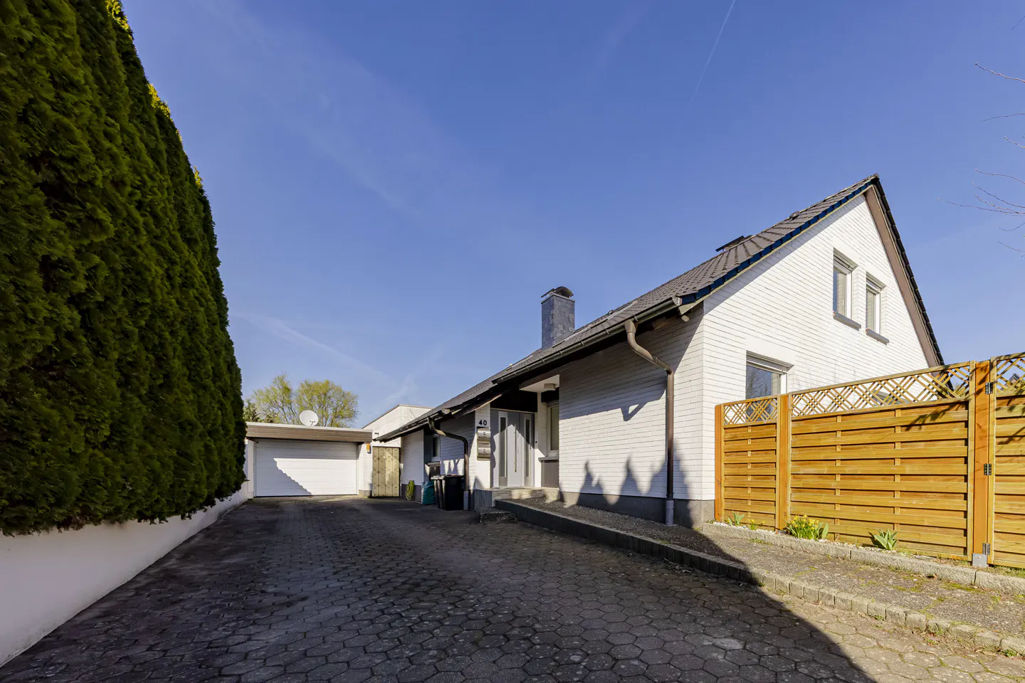Exterior view of a white, one-story house with a dark roof, chimney, and a paved driveway under a blue sky. A tall green hedge borders the left side.