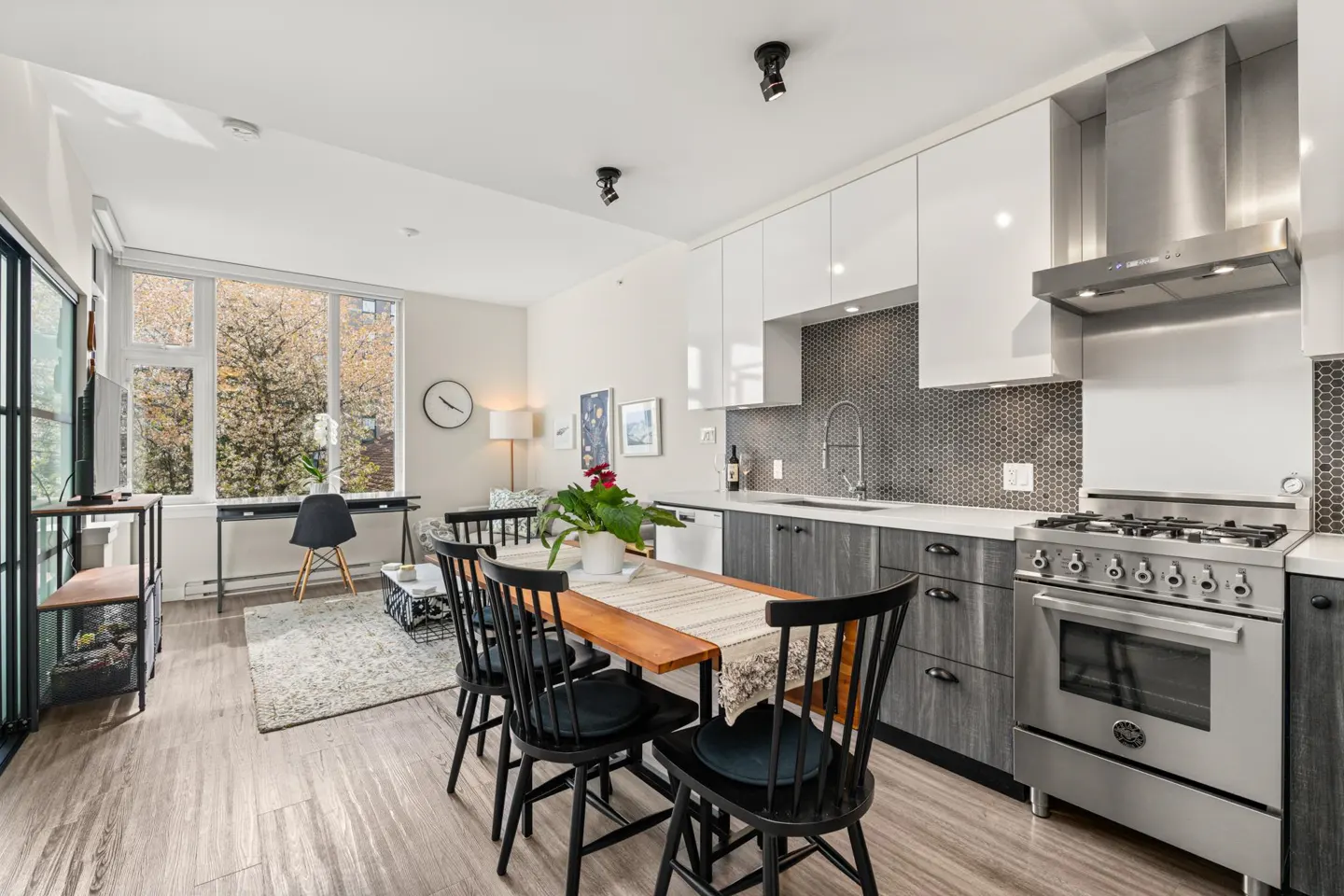 Bright, open-concept apartment with a kitchen featuring white cabinets, stainless steel appliances, and a wooden dining table with black chairs.