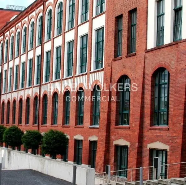 Exterior of a red brick commercial building with arched windows and green trim. A row of green bushes lines the base of the building.