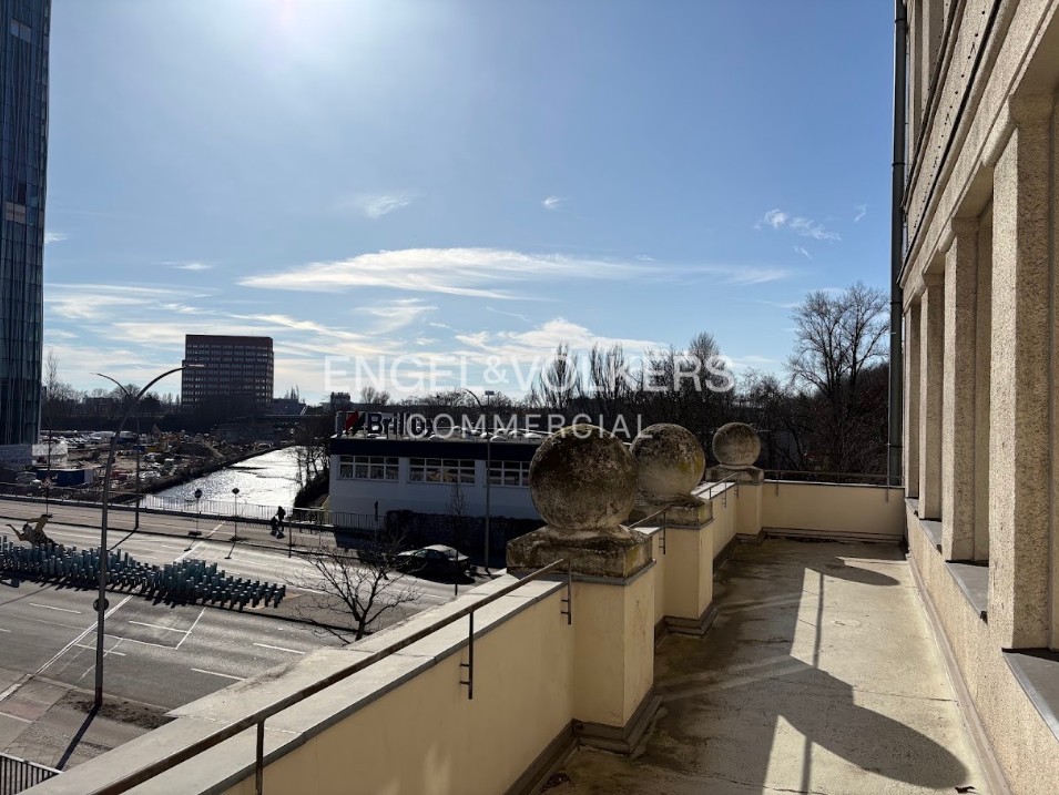 View from a beige balcony with stone spheres overlooking a city street, a canal, and distant buildings under a blue sky.