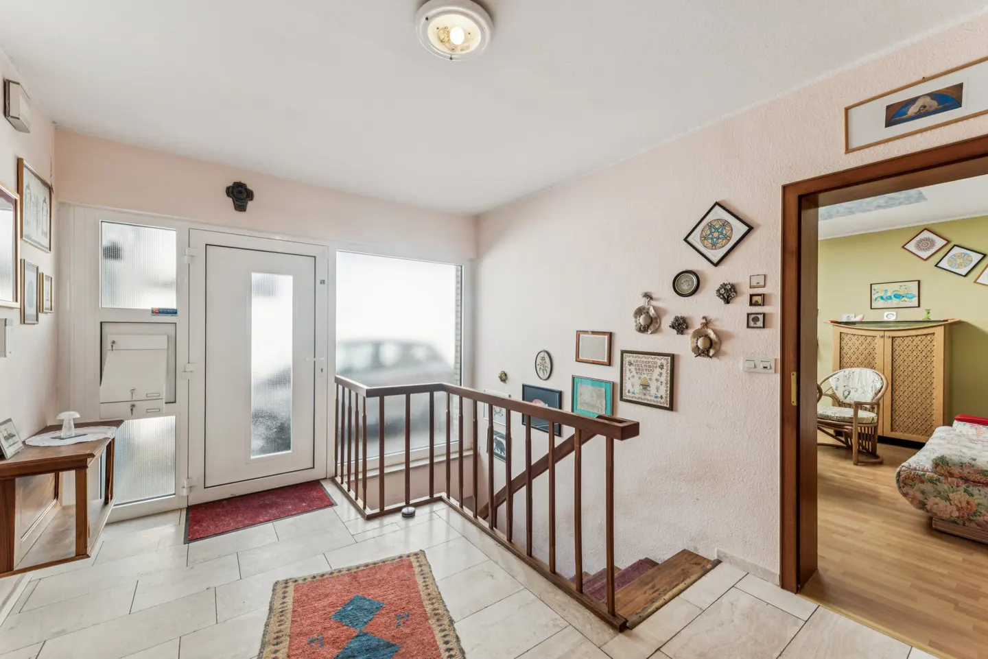 Entryway with white tile floor, a white door, and a wooden staircase with brown railing. Walls are light pink, decorated with framed pictures. A glimpse of a living room is visible.