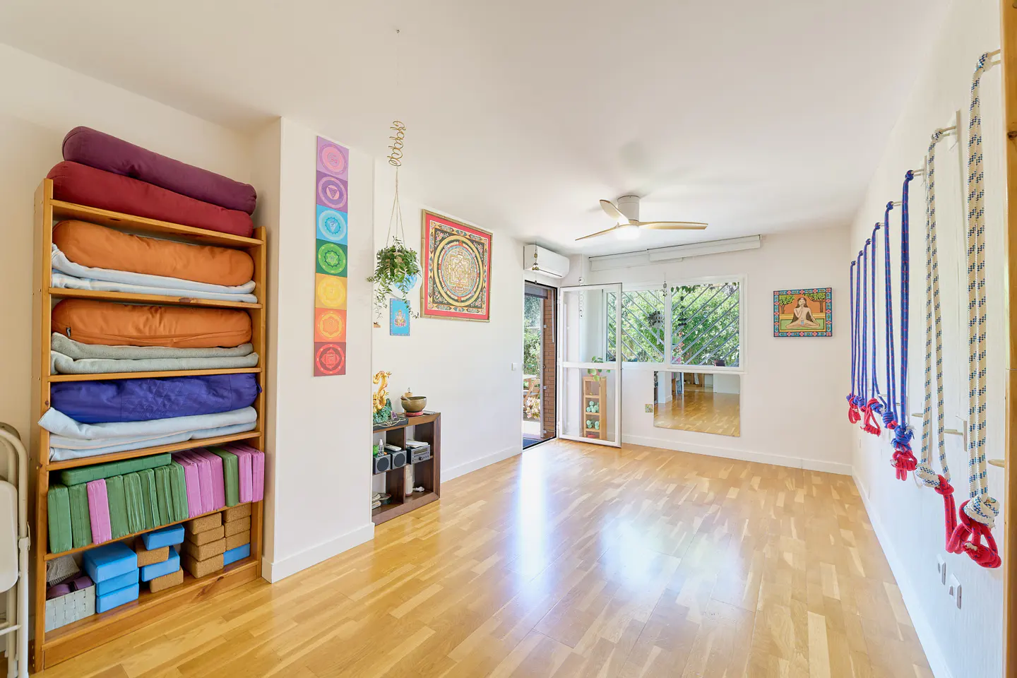 Bright yoga studio with wood floors, white walls, and natural light. Shelves hold colorful bolsters and blocks. Ropes hang on the right wall.