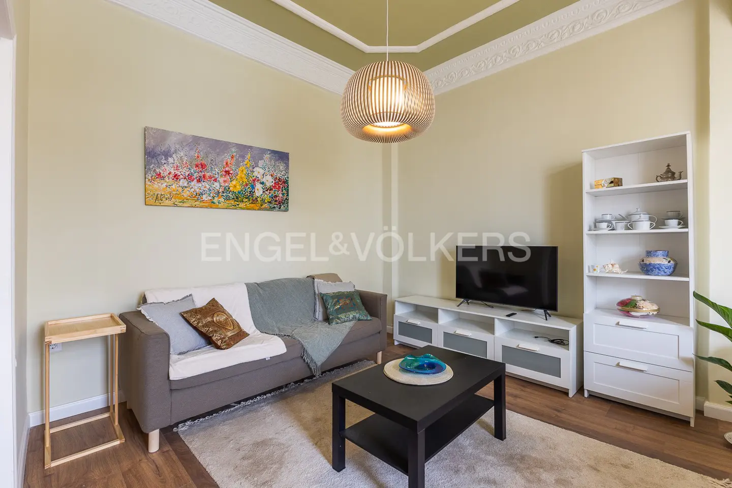 Living room with a gray sofa, black coffee table, TV, and white shelving unit. A round, modern light fixture hangs from the ceiling.