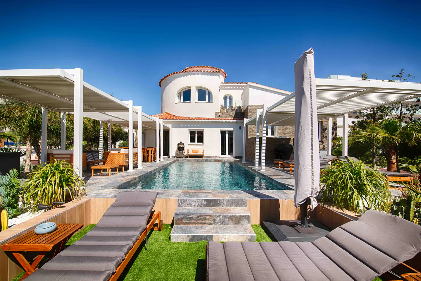 Luxury home exterior with a pool, lounge chairs, and white pergolas under a clear blue sky. The house has a round tower and a red tile roof.