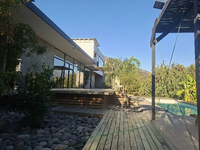 Exterior view of a modern house with a wooden deck, pool, and lush greenery against a clear blue sky.