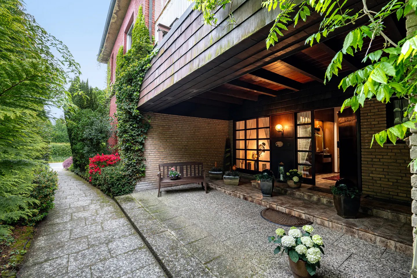 Exterior view of a brick house with a covered porch, stone walkway, bench, and lush greenery. The entrance has glass doors and warm lighting.