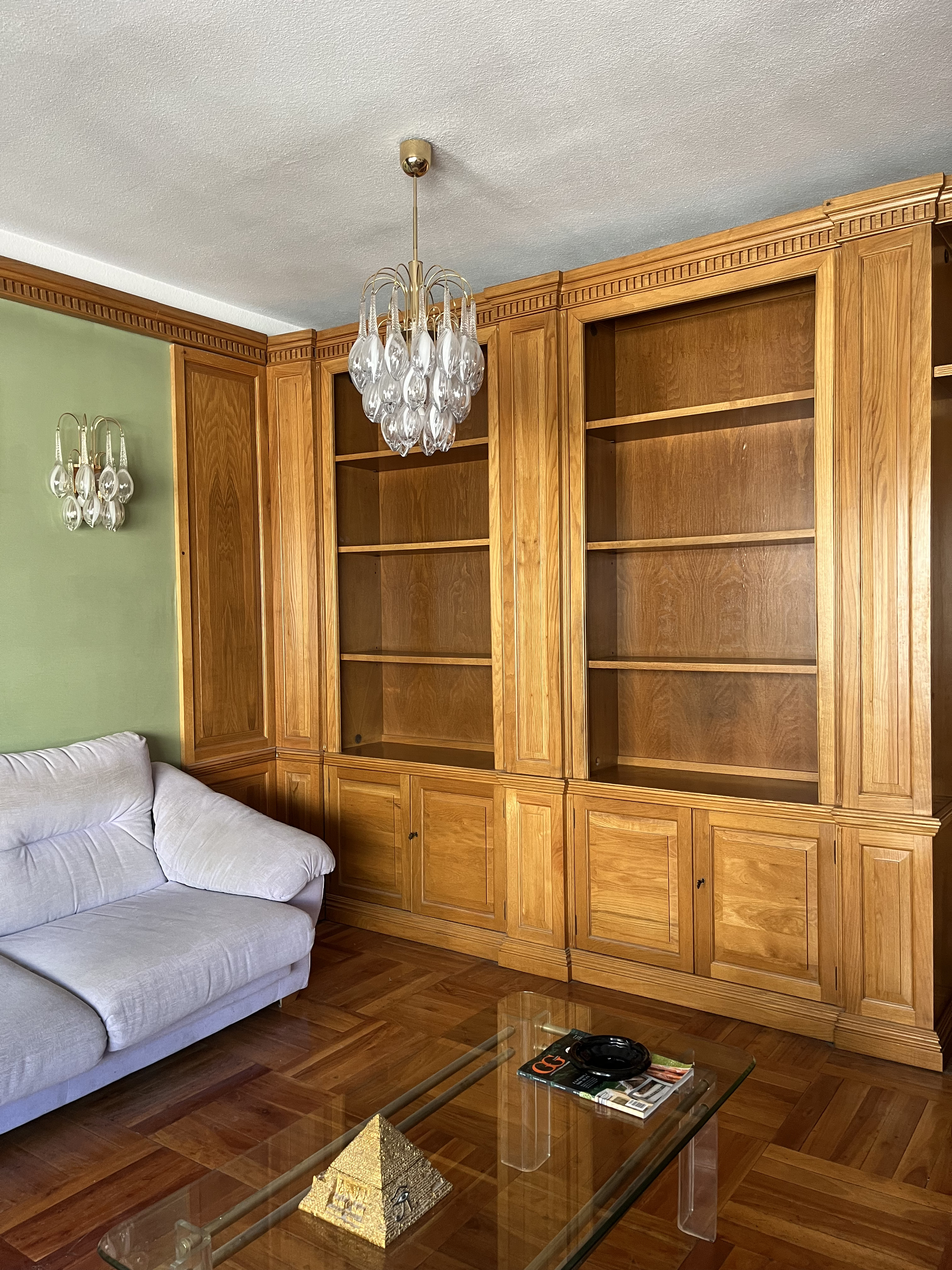 Living room with a light grey sofa, large wooden built-in bookshelves, a glass coffee table, and matching crystal chandeliers. [AI-generated text]