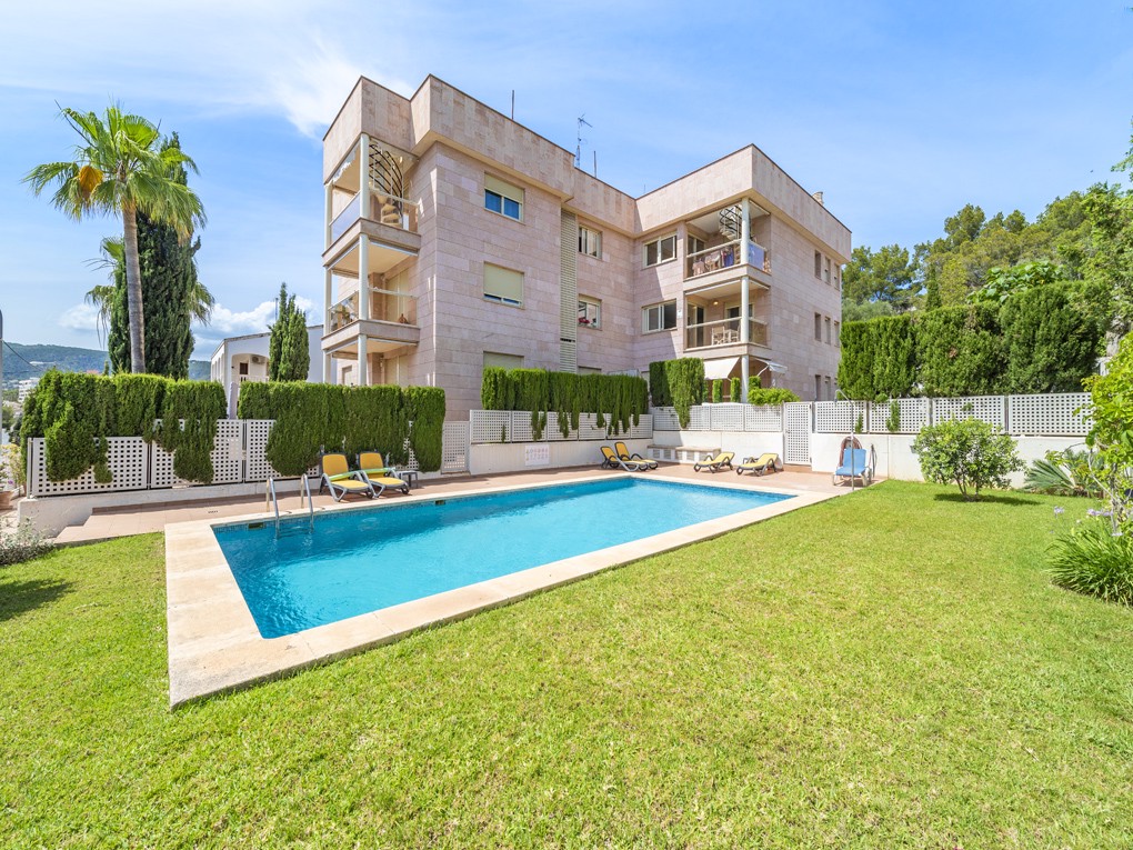 A multi-story stone apartment building with a swimming pool, green lawn, and lounge chairs under a clear blue sky.