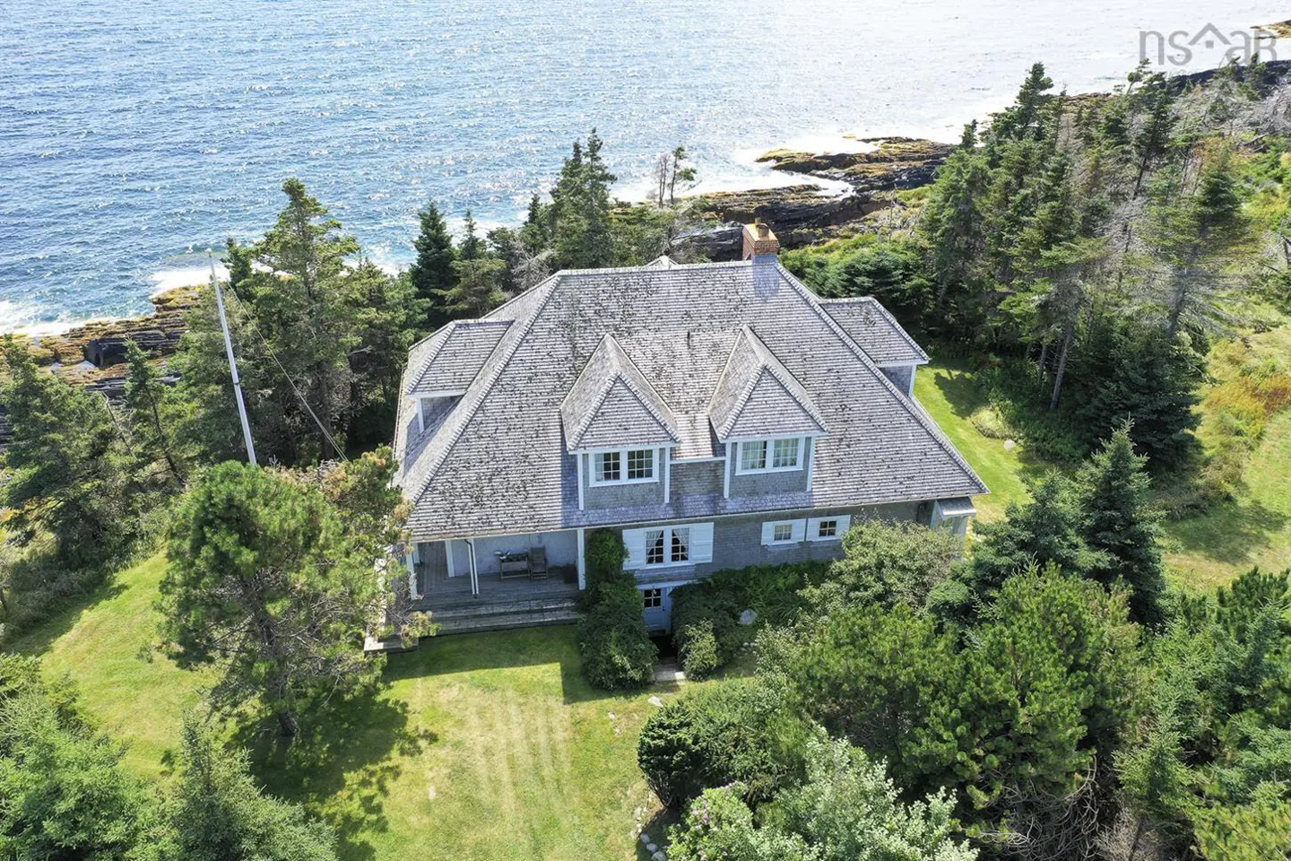 Aerial view of a grey shingle-style house with white trim, nestled amongst green trees near the blue ocean.