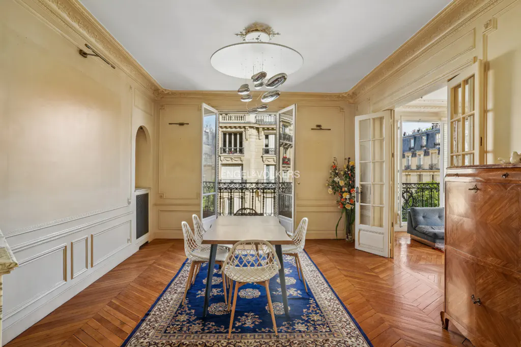 A bright dining room with a table, chairs, and open doors to a balcony overlooking a city. The room has wood floors and a blue rug.