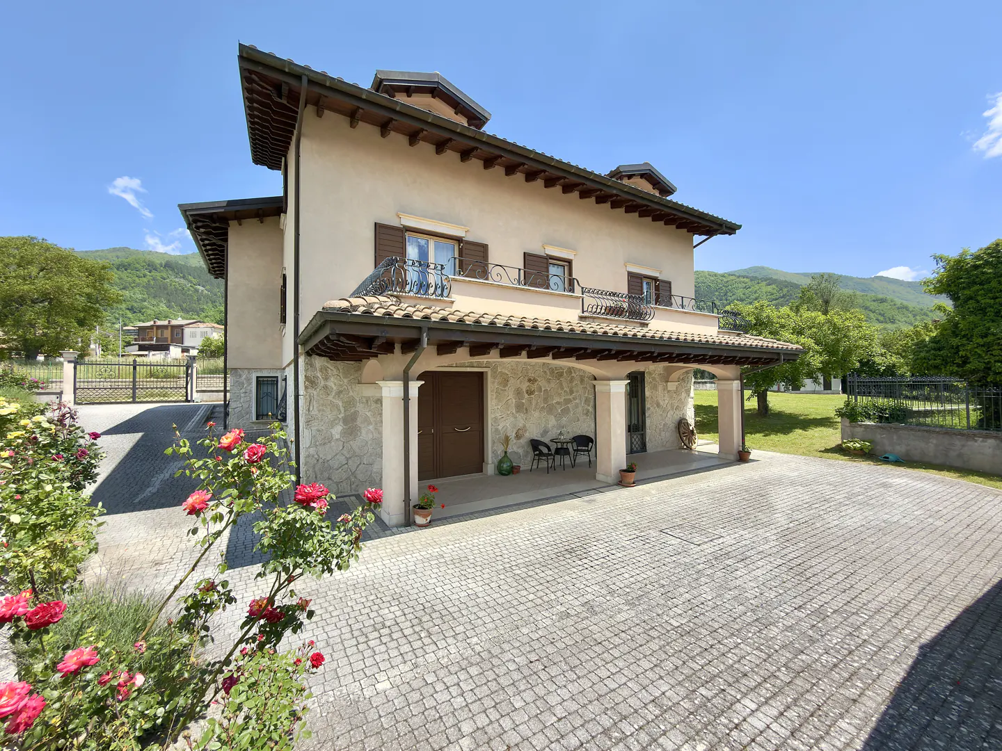 Two-story beige villa with stone facade, covered patio, and cobblestone driveway, set against a backdrop of green mountains.
