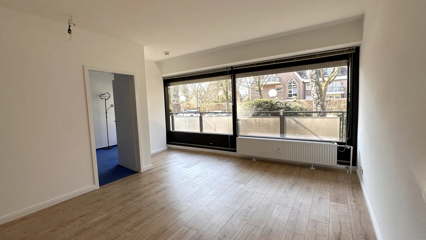 Bright, empty room with wood floors, white walls, and a large window overlooking a balcony and street. A doorway leads to a room with blue carpet.