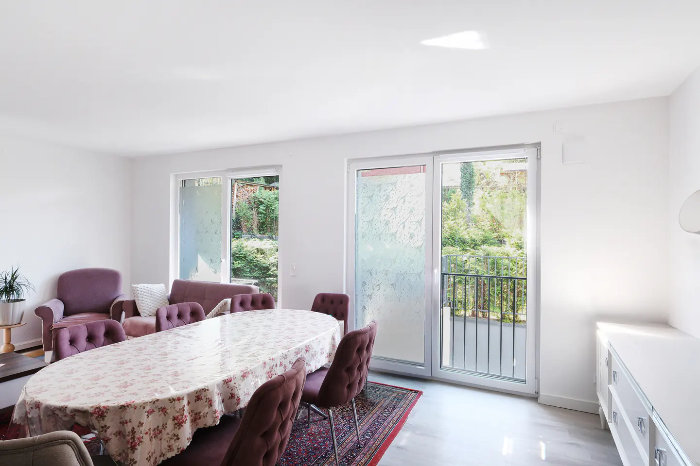 Bright dining room with white walls, a floral tablecloth on an oval table, and purple chairs and sofas. Sliding glass doors lead to a balcony.