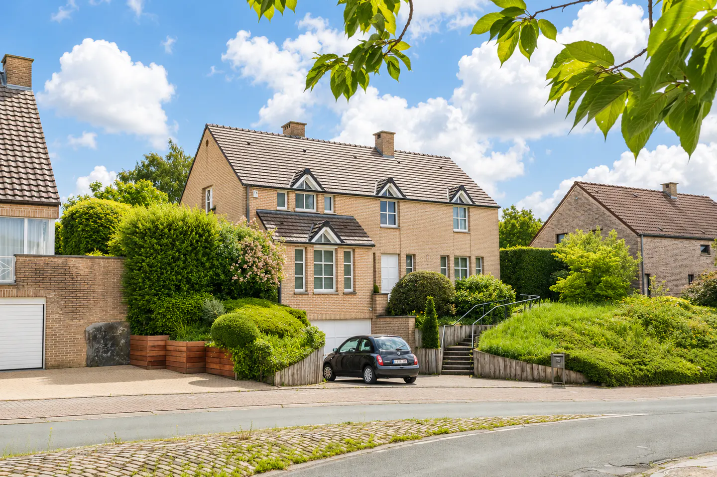 A two-story tan brick house with a brown tiled roof, lush landscaping, and a black car parked in the driveway.