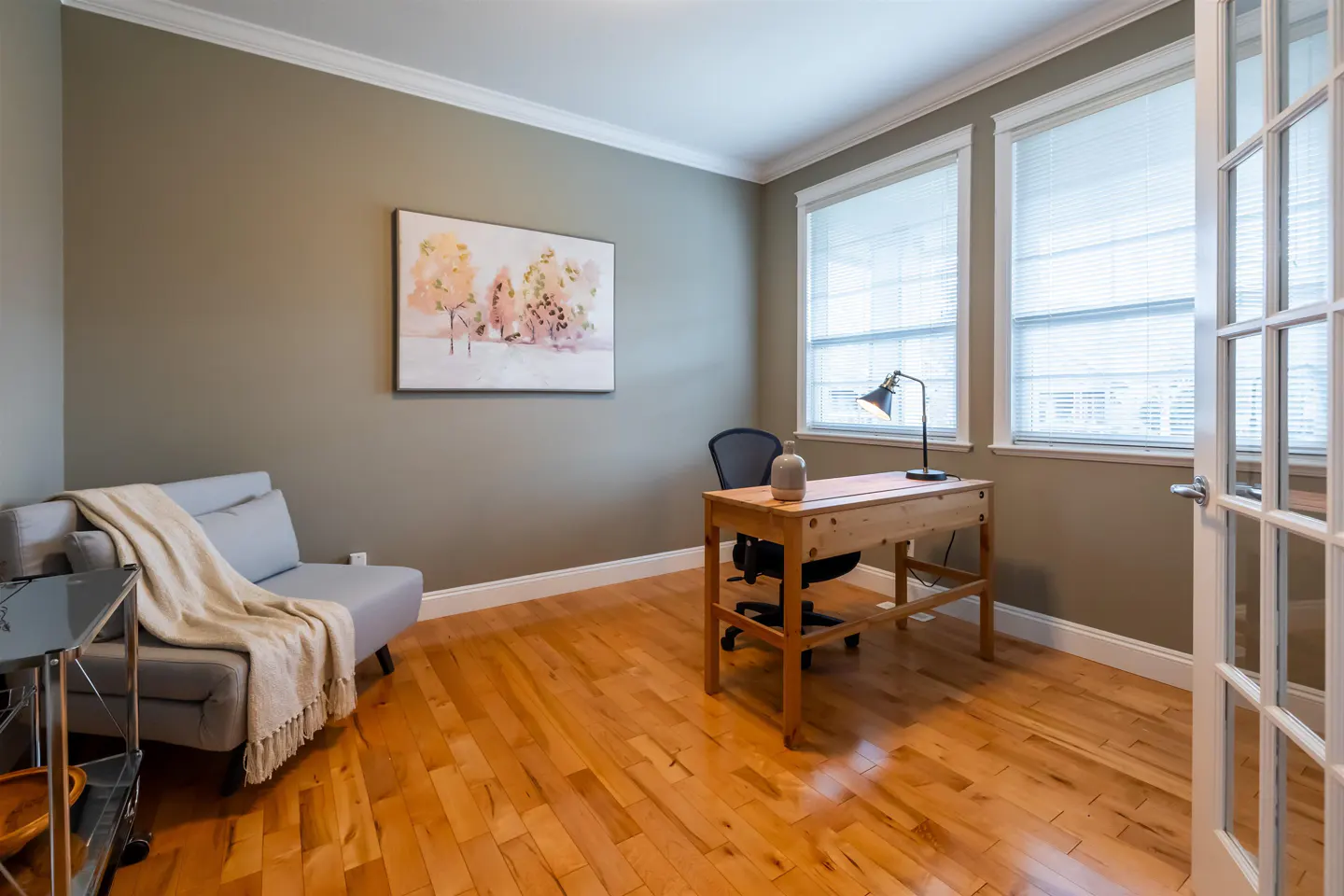 A home office with hardwood floors, a gray sofa with a throw, a desk, and a chair. Two windows with blinds let in natural light.