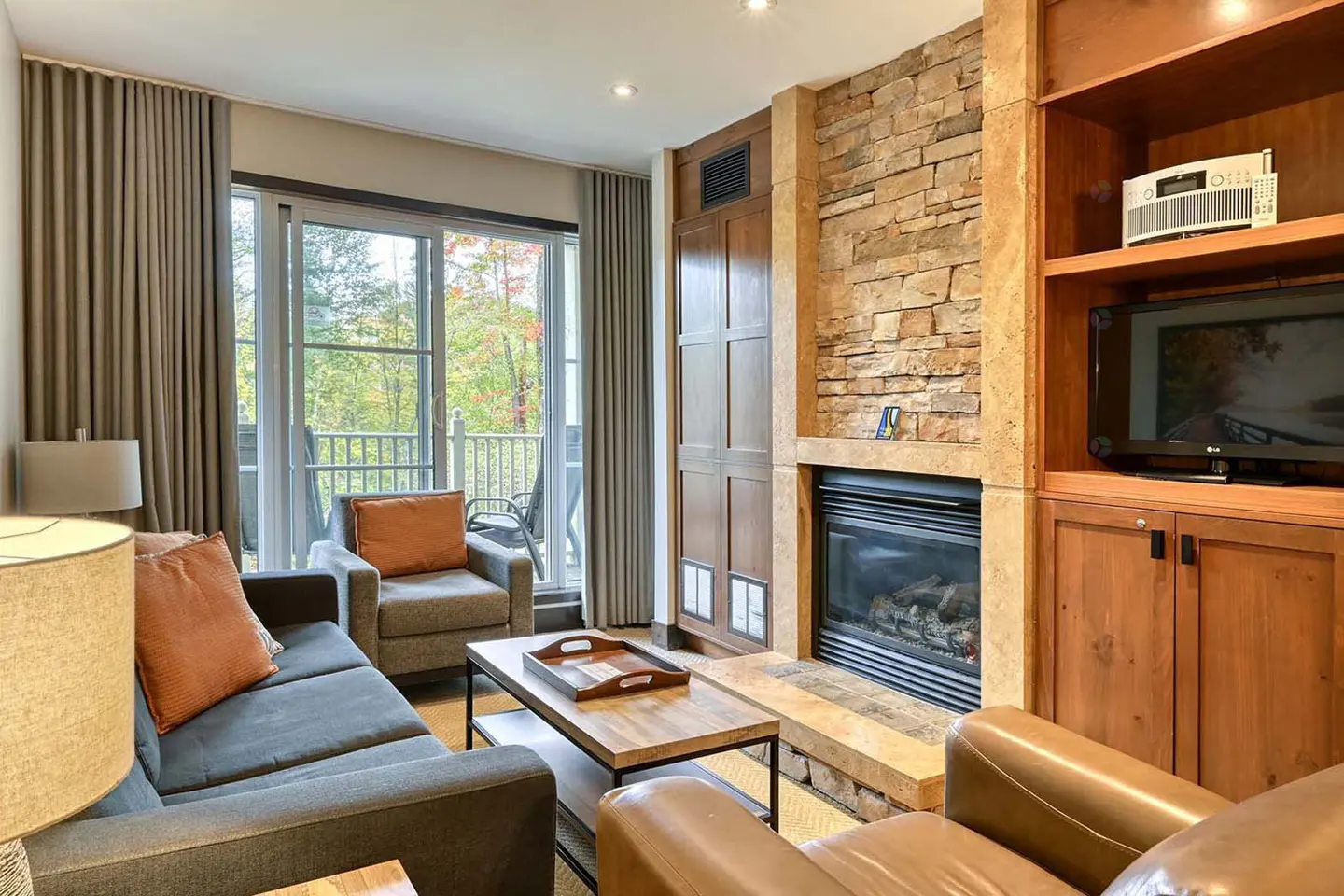 Living room with gray sofa, leather chair, stone fireplace, and balcony with trees in the background.