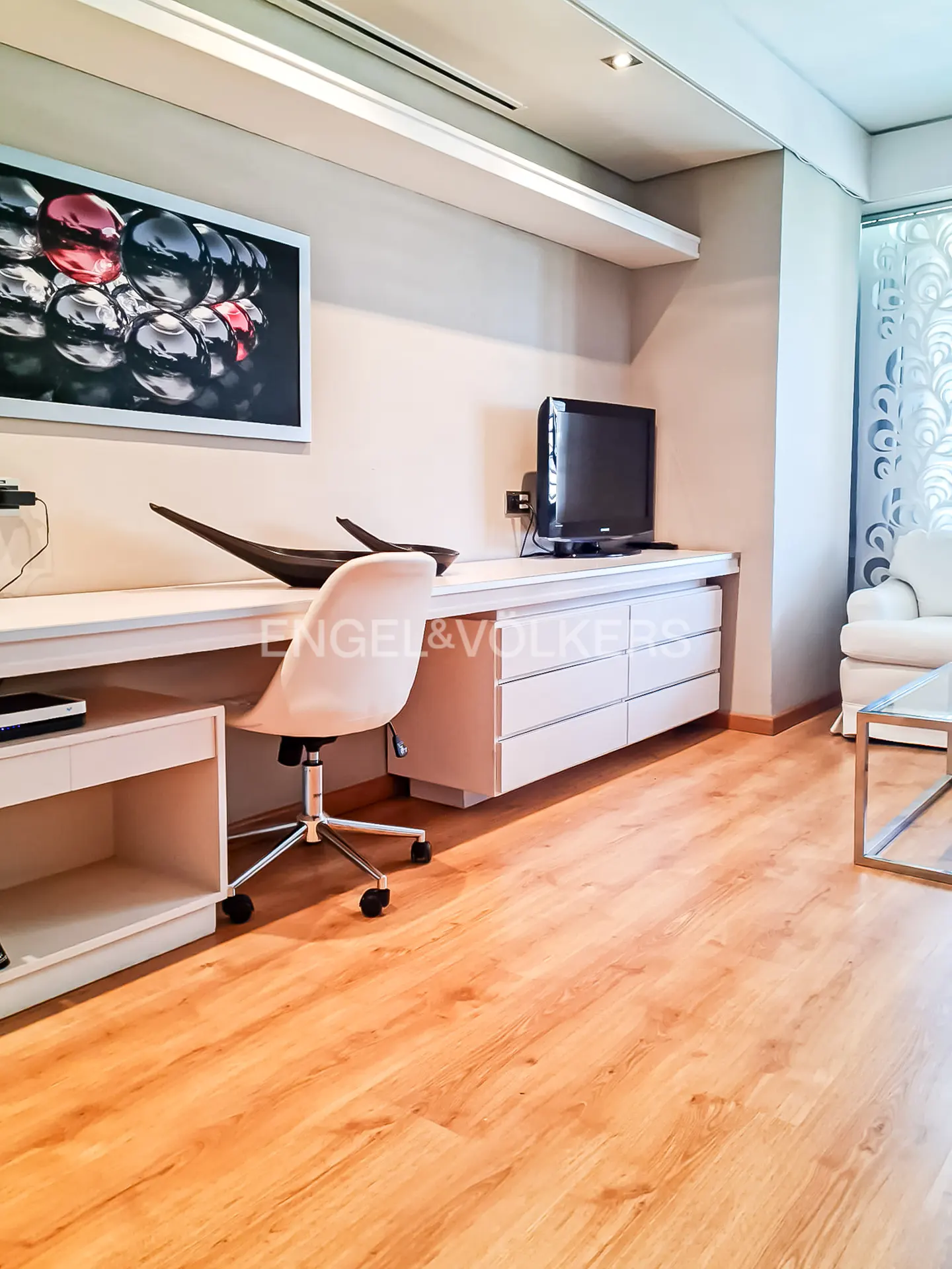 Modern home office with a white desk, swivel chair, television, and abstract wall art on light wood flooring.